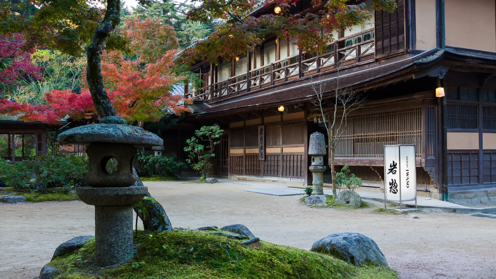 A traditional Japanese ryokan entrance with autumn maples in red and orange beside a stone lantern and moss-covered garden