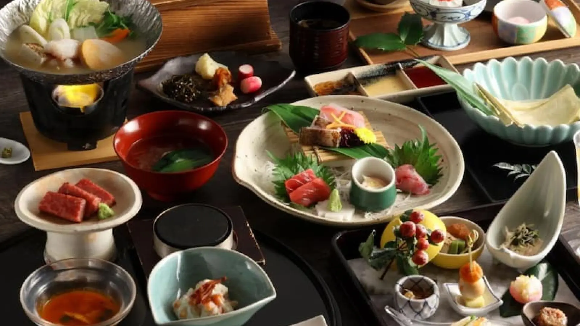 A full kaiseki dinner spread across a dark lacquer table, featuring sashimi, simmered dishes, grilled fish, and seasonal garnishes on varied ceramics