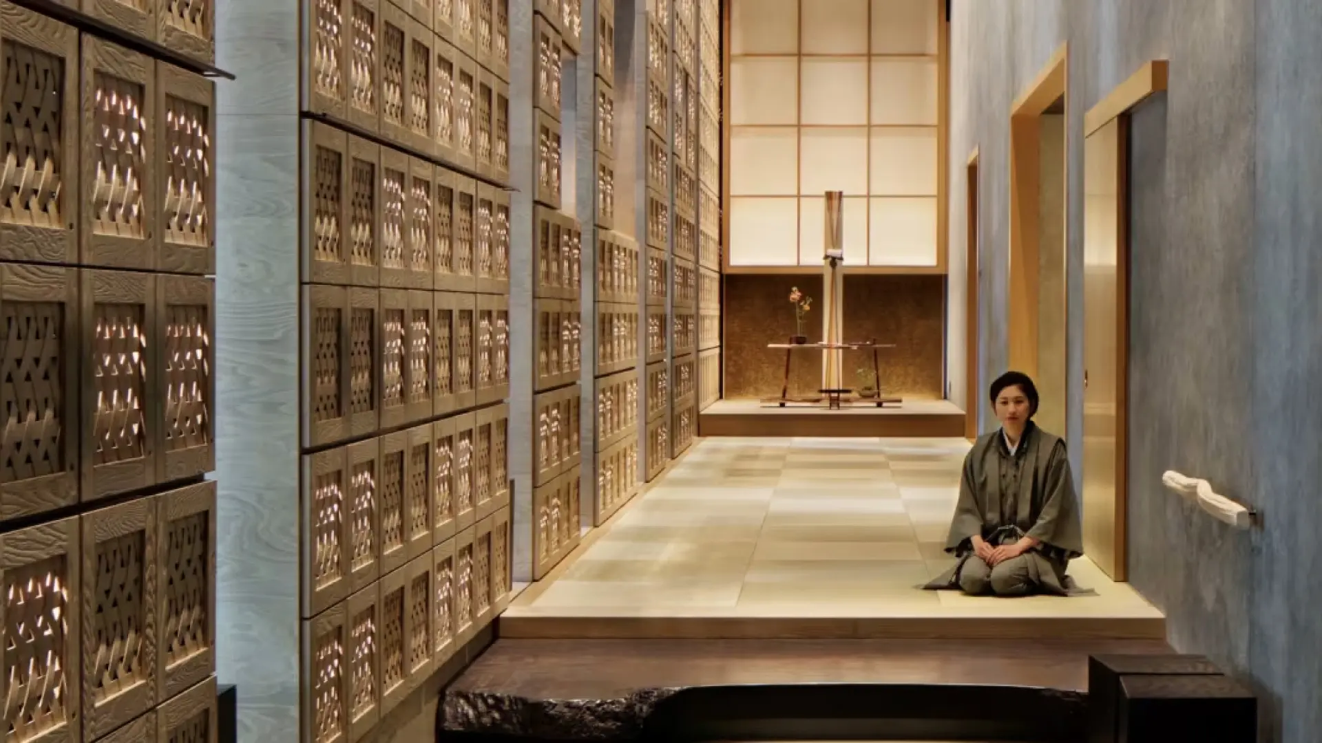 A ryokan concierge in dark kimono seated on a tatami platform in a modern hallway lined with traditional wooden cabinets