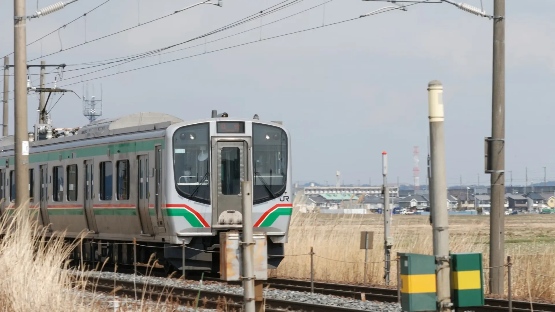 A JR regional train with green and red striping approaching on rural tracks under an overcast sky