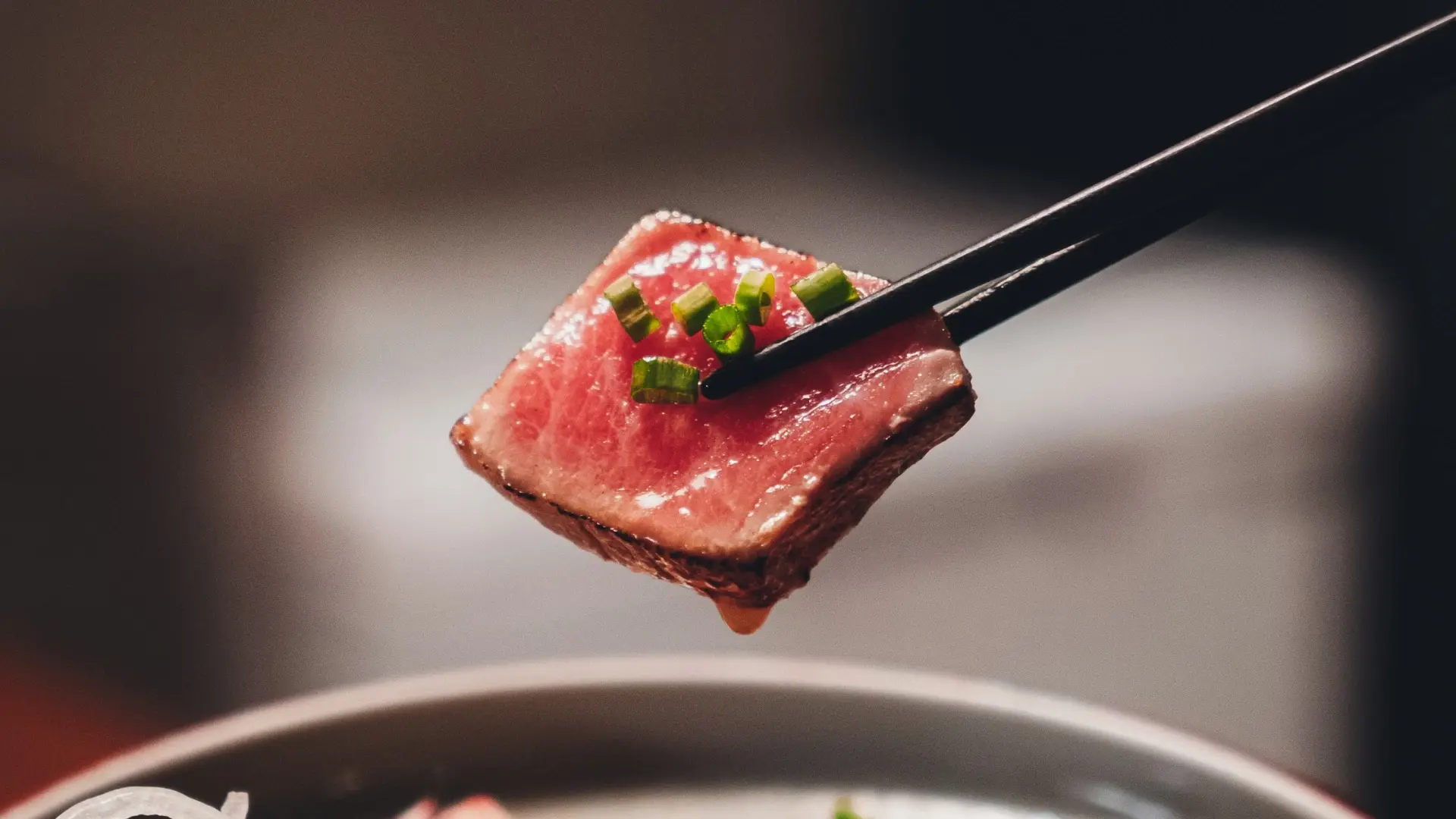A slice of seared wagyu beef with chopped scallions held by black chopsticks above a steaming pot