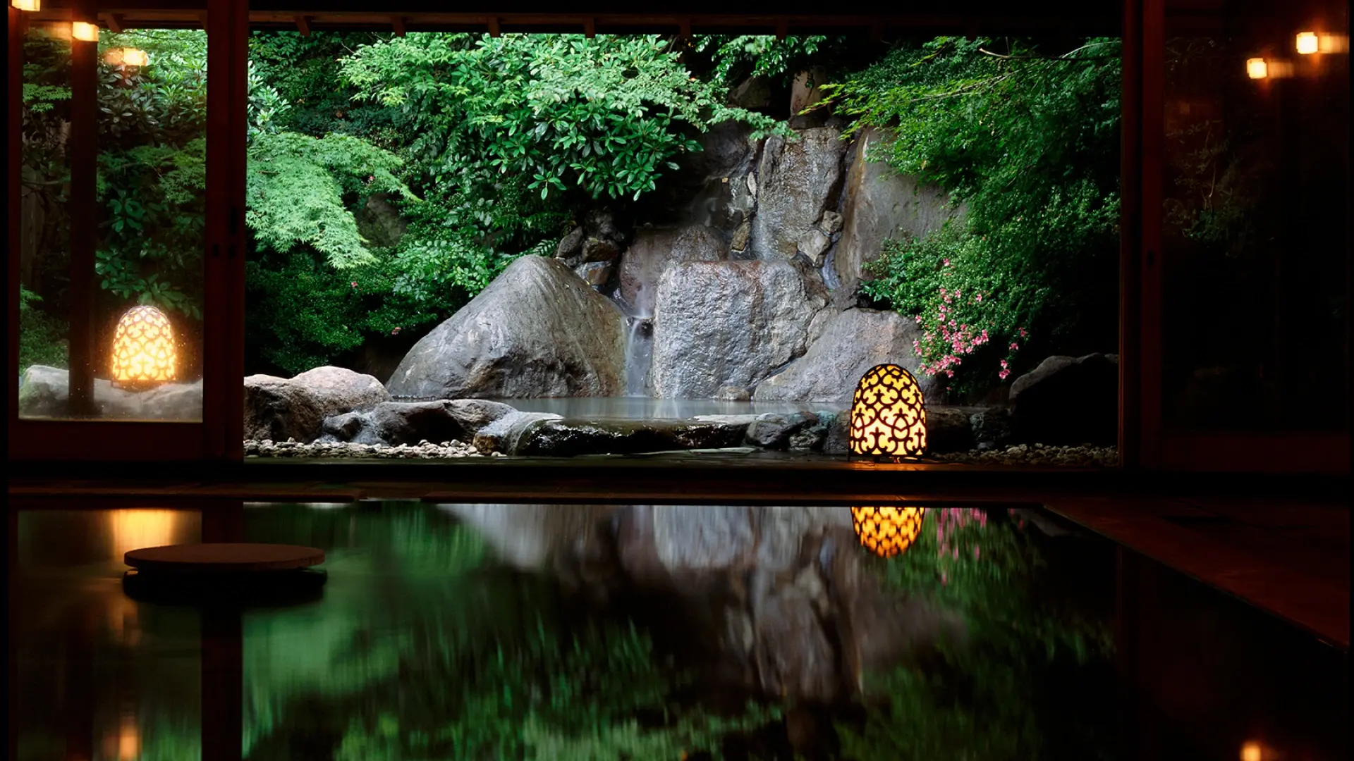 An indoor onsen bath at night with illuminated garden boulders and glowing lanterns reflected in the still water