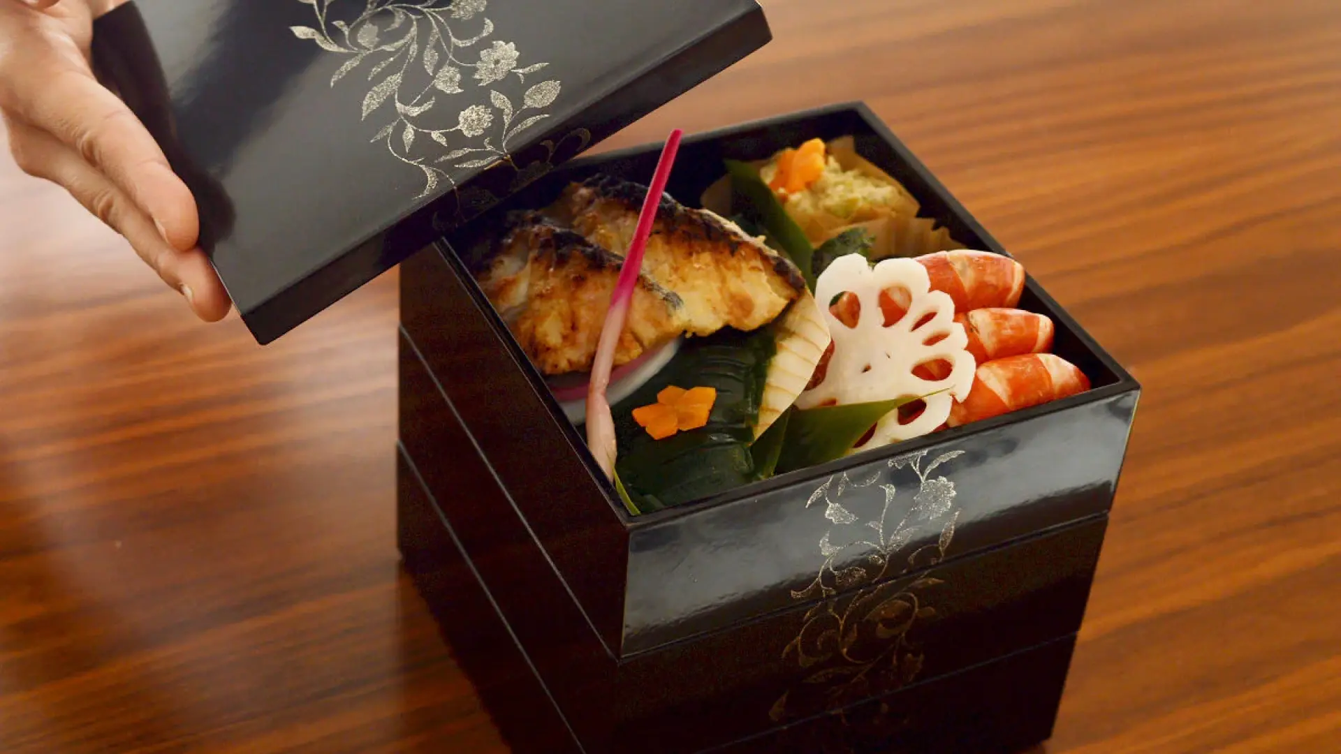 A hand lifting the lacquered lid of a jubako tiered box revealing osechi ryori New Year dishes with lotus root, grilled fish, and colorful garnishes