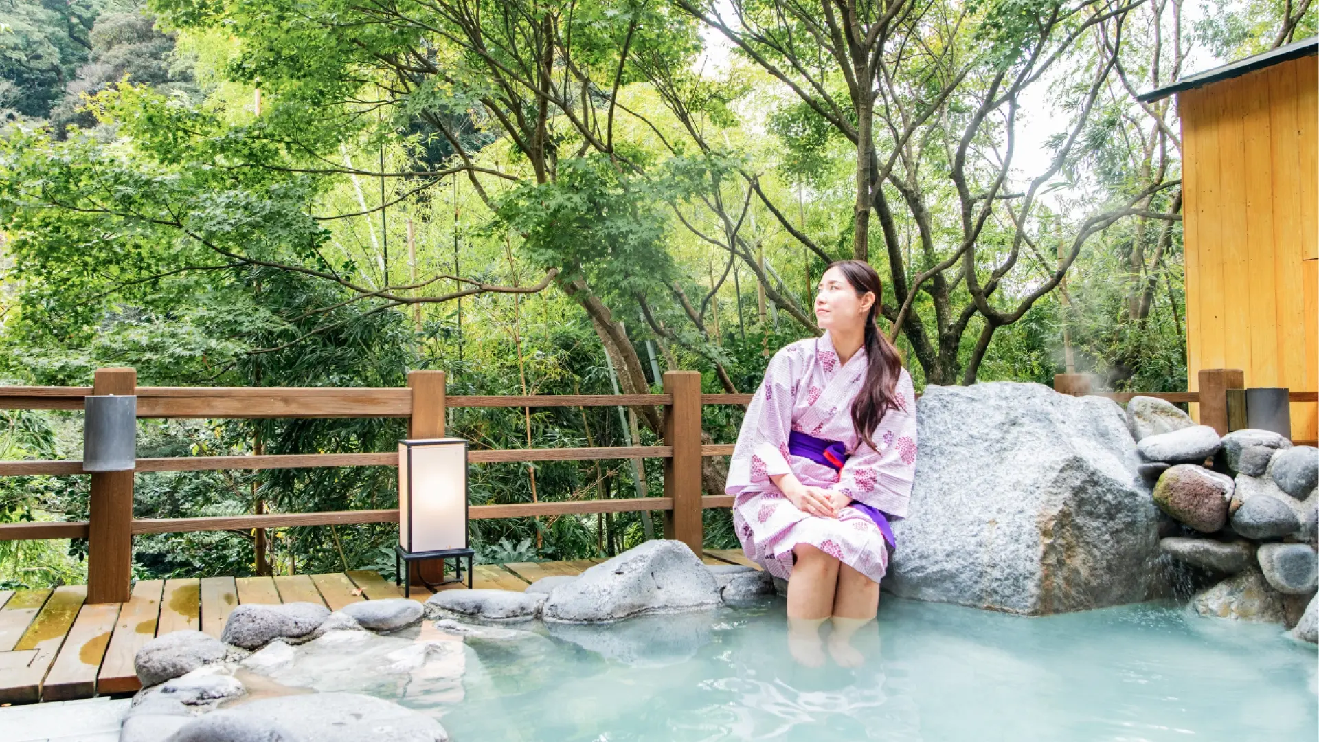 Woman in pink yukata sitting at the edge of an open-air stone onsen bath surrounded by lush green forest