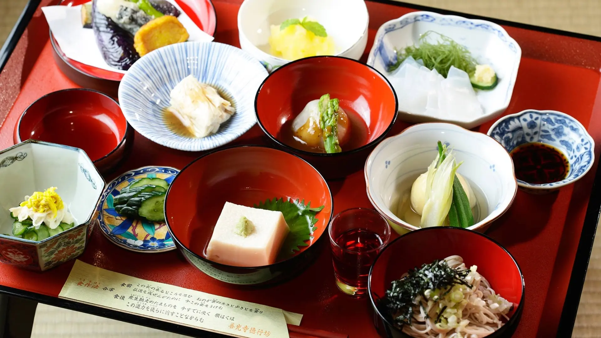 A shojin ryori vegetarian meal arranged on a red lacquer tray with tofu, simmered vegetables, pickles, and soba noodles in individual bowls