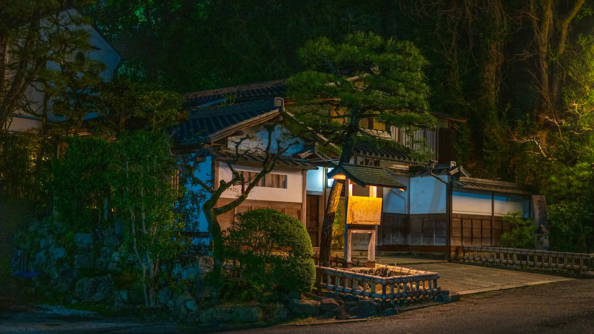 A traditional ryokan entrance illuminated by a warm noren curtain at dusk, surrounded by sculpted pines and stone walls