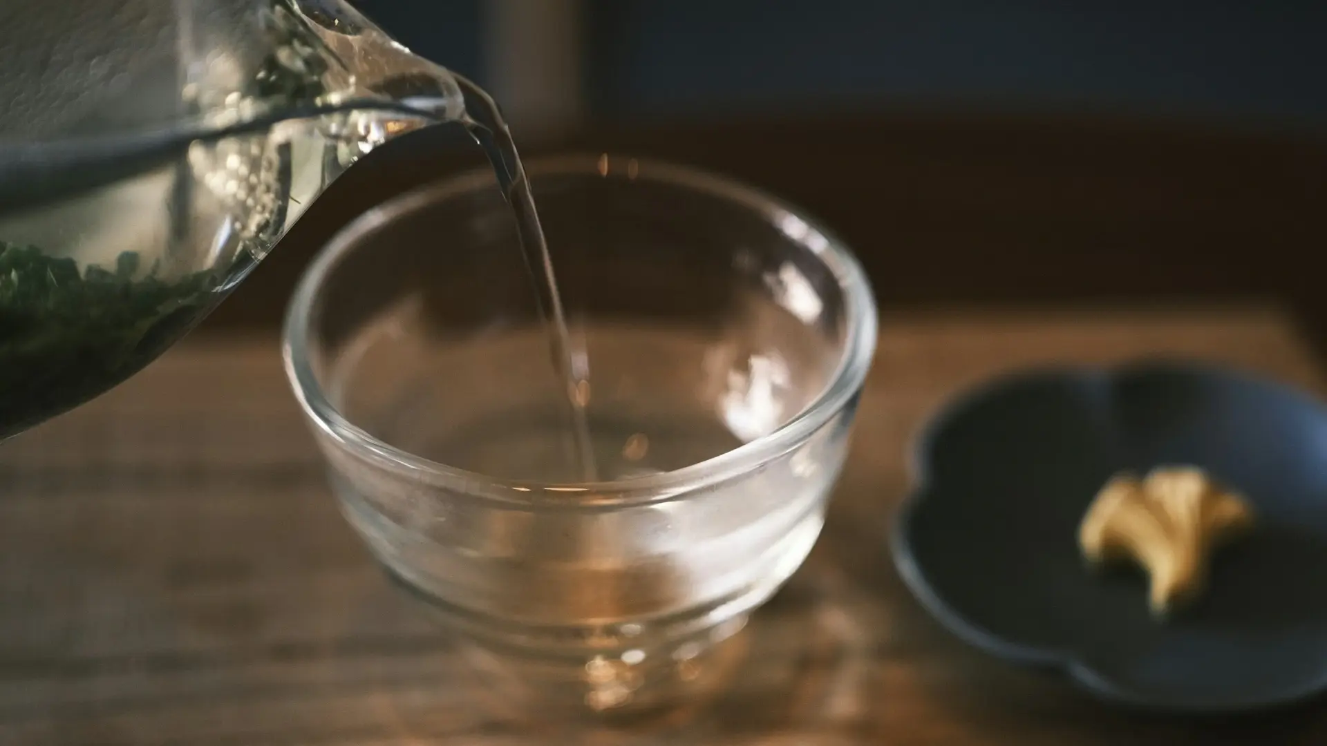 Green tea being poured from a glass teapot into a clear glass bowl, with a small wagashi confection on a dark plate beside it