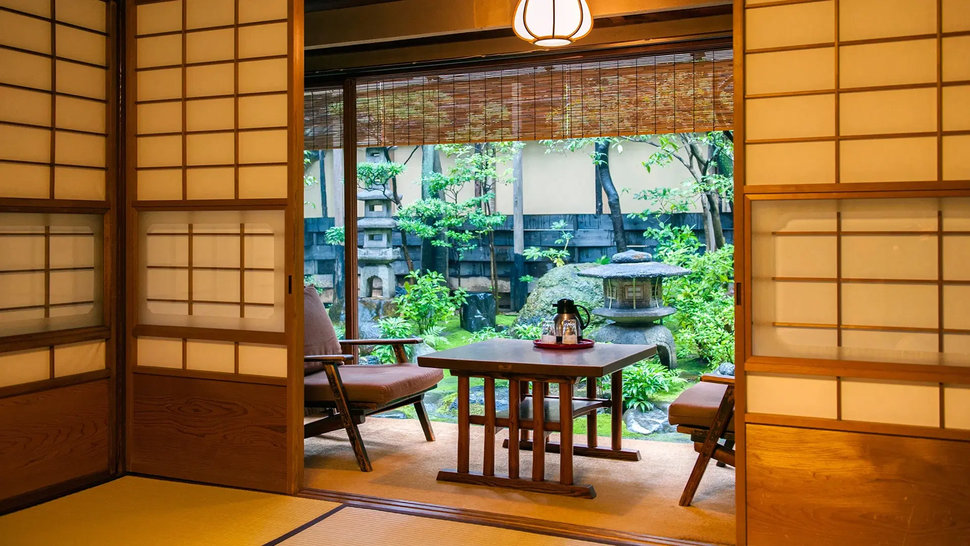 Ryokan sitting room with low wooden table and chairs framed by shoji screens, looking onto a lush Japanese garden with stone lanterns