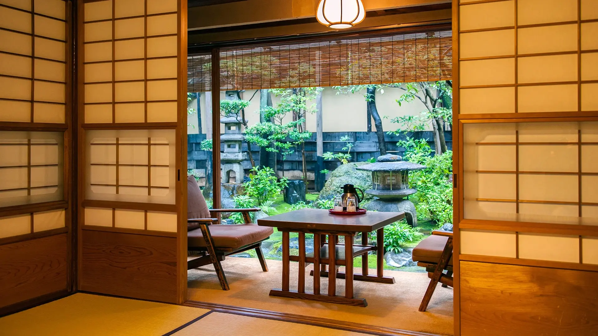 Ryokan sitting room with low wooden table and chairs framed by shoji screens, looking onto a lush Japanese garden with stone lanterns