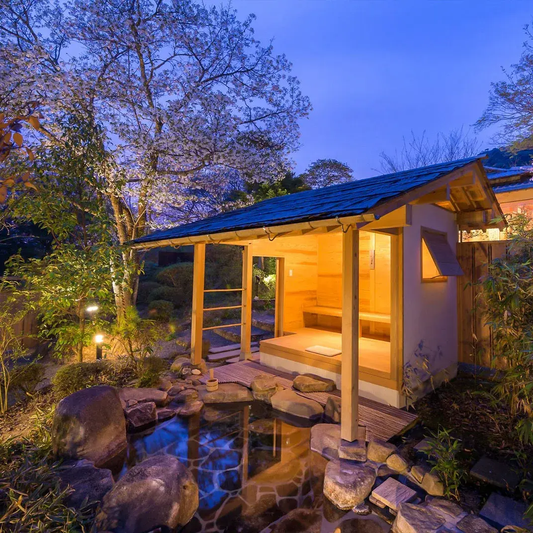 Private rotenburo glowing beneath lit cherry blossoms and stone lanterns at dusk