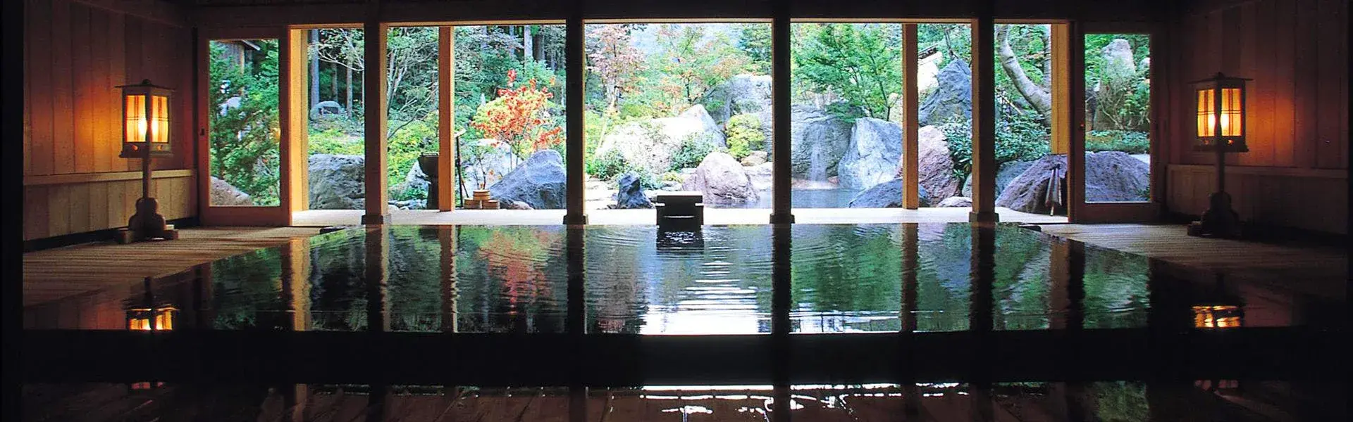 Indoor onsen bath reflecting lantern light across still water toward a rock garden