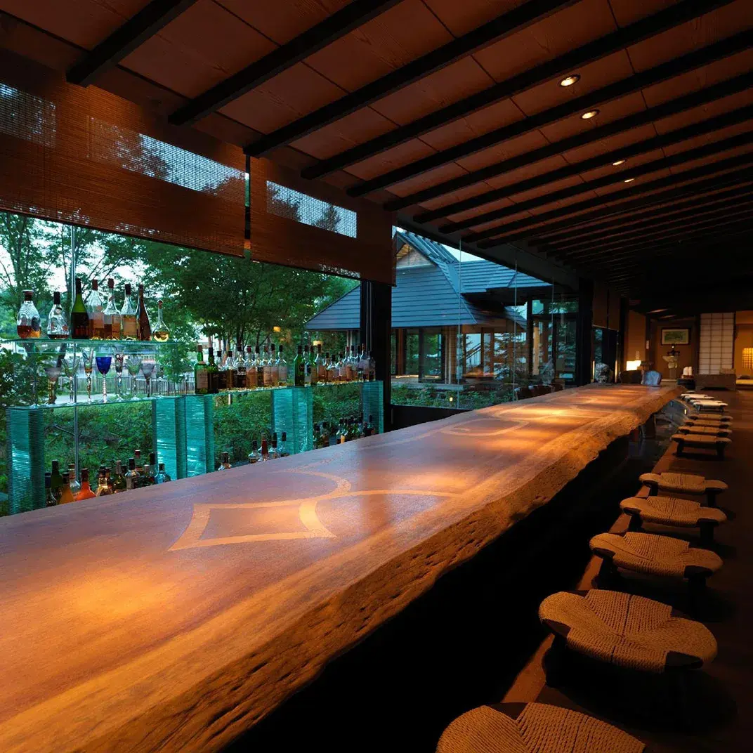 Live-edge wood bar counter lit at dusk, liquor bottles lining the garden-facing window