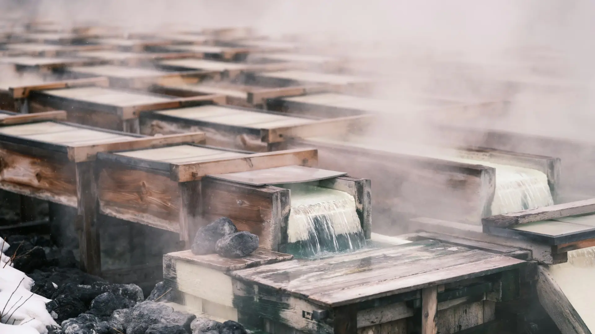 Rows of wooden yubatake boxes channeling steaming hot spring water with mineral deposits, shrouded in thick vapor