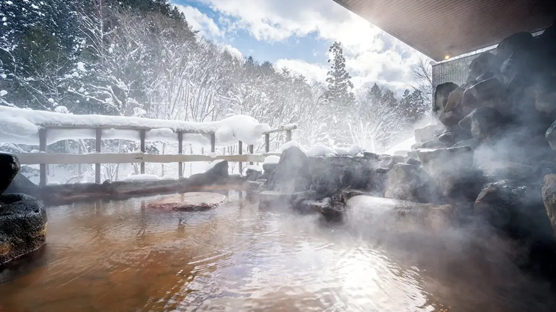 A steaming outdoor rotenburo bath with iron-tinted water surrounded by snow-covered rocks and bare winter trees