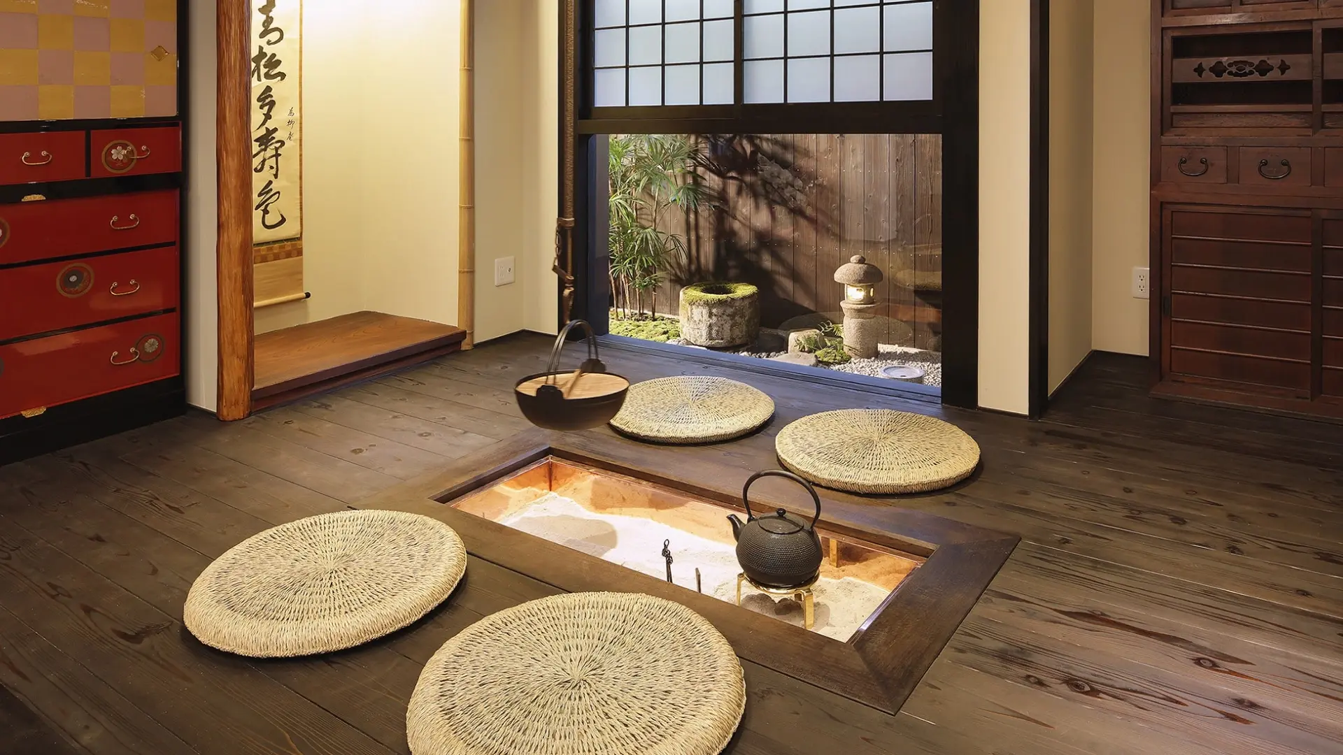 Traditional irori hearth set into dark wooden floorboards with woven zabuton cushions, a hanging kettle, and a courtyard garden beyond