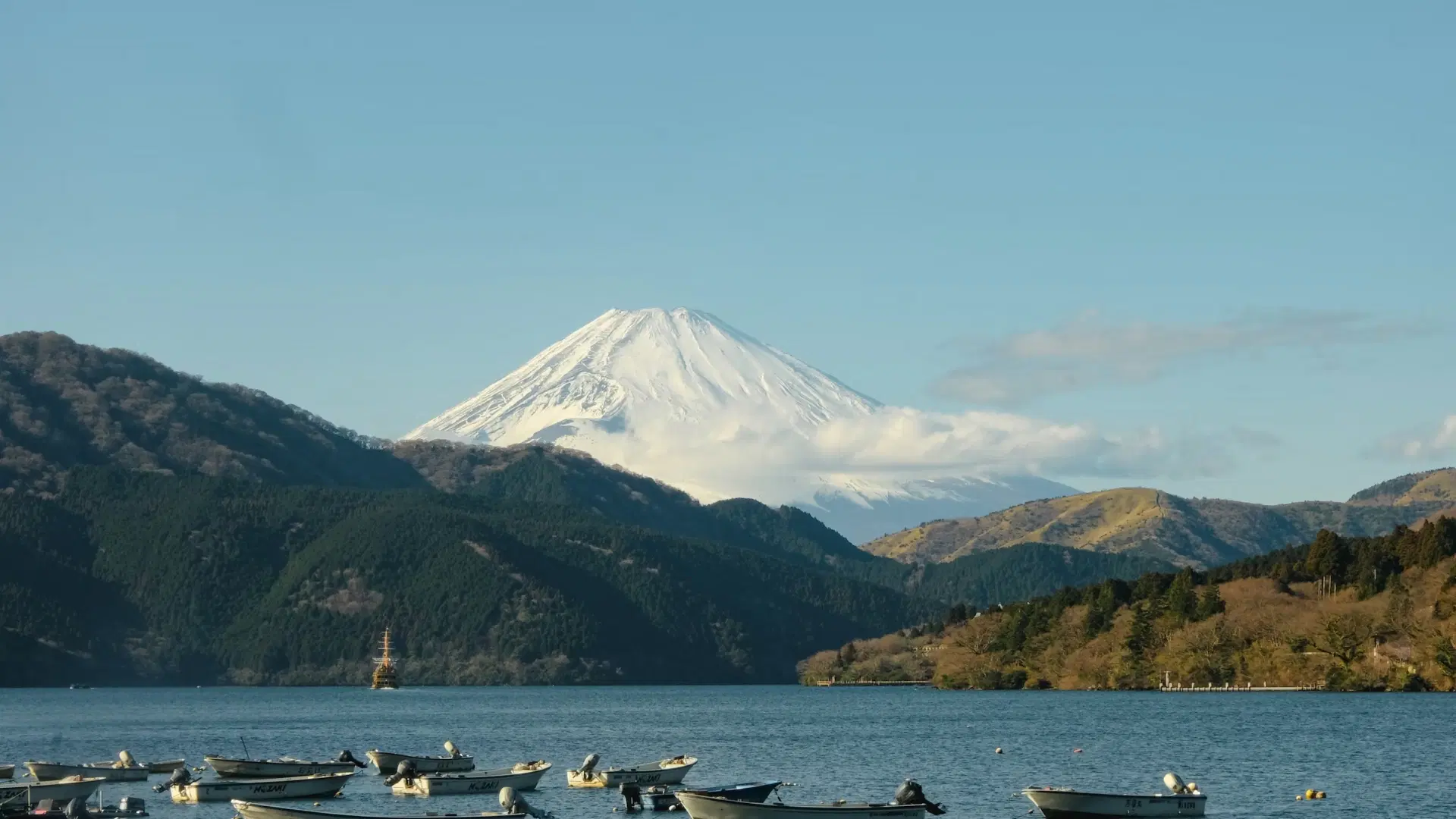 Snow-capped Mount Fuji rising above forested hills and Lake Ashi with small boats moored in the foreground
