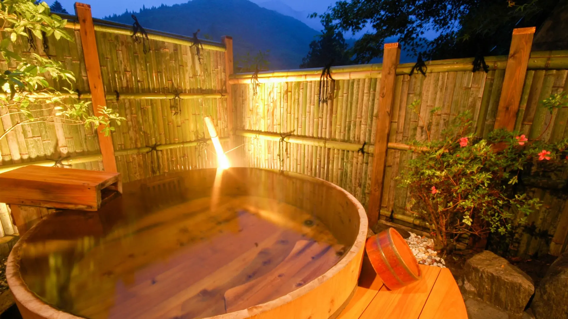A private wooden barrel bath steaming at dusk, enclosed by a bamboo fence with mountains visible beyond