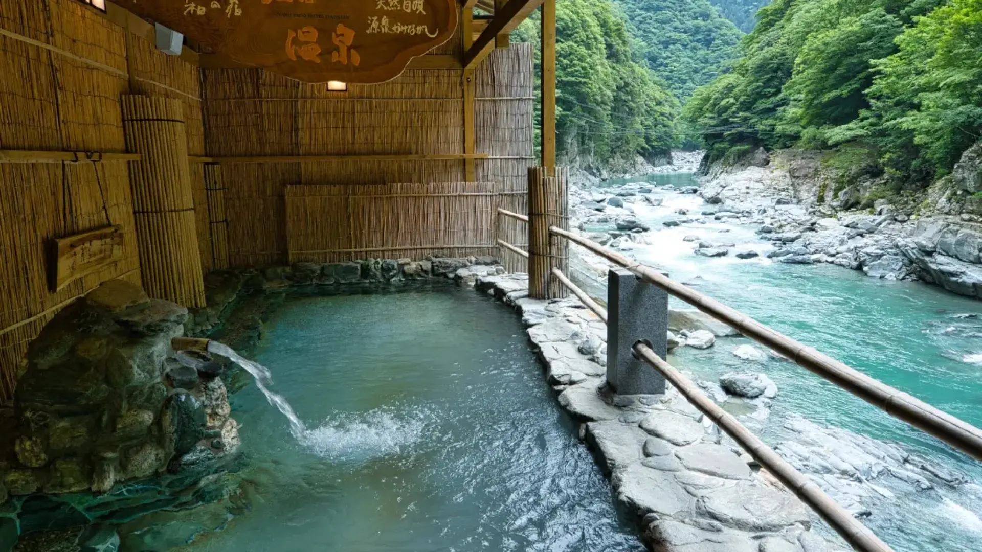 A riverside rotenburo with milky blue water beside a rushing mountain river, sheltered by a bamboo screen and lush green canopy
