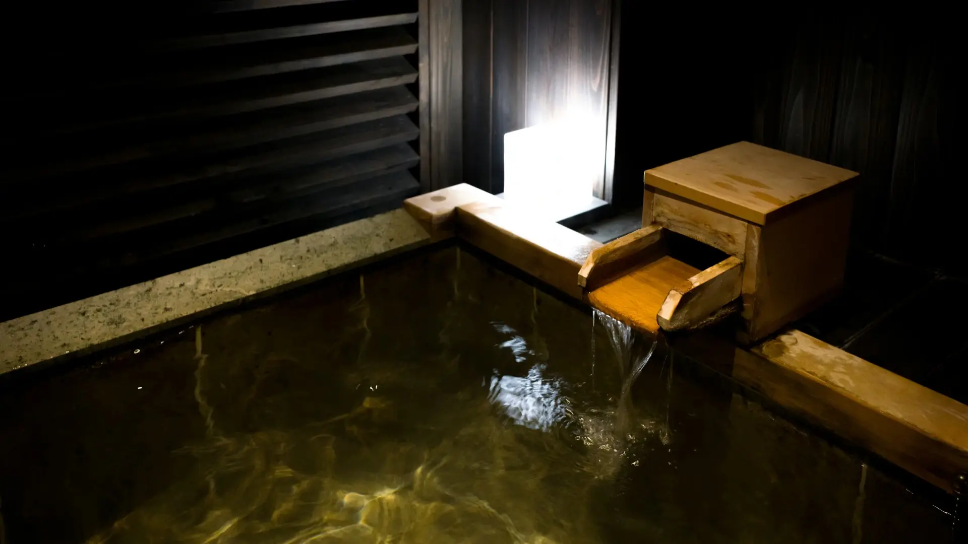 Hot spring water flowing from a wooden kakei spout into a stone-edged indoor bath with amber-tinted water