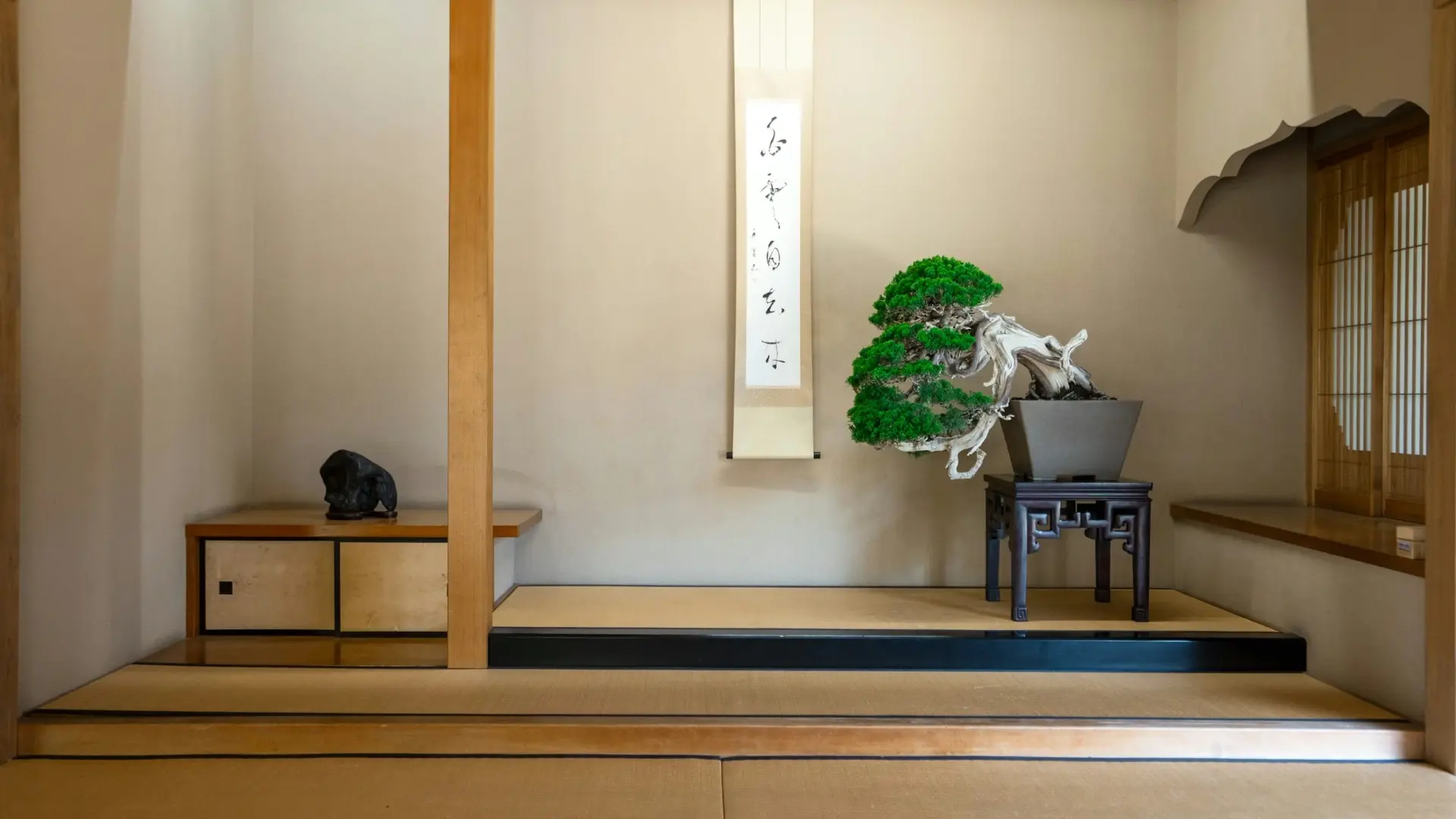 A tokonoma alcove displaying a hanging calligraphy scroll and a sculpted bonsai pine on a dark wooden stand