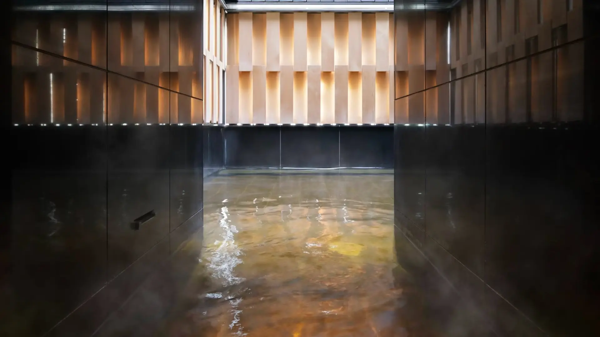 A modern indoor onsen bath with amber-tinted mineral water, steam rising beneath soft overhead lighting